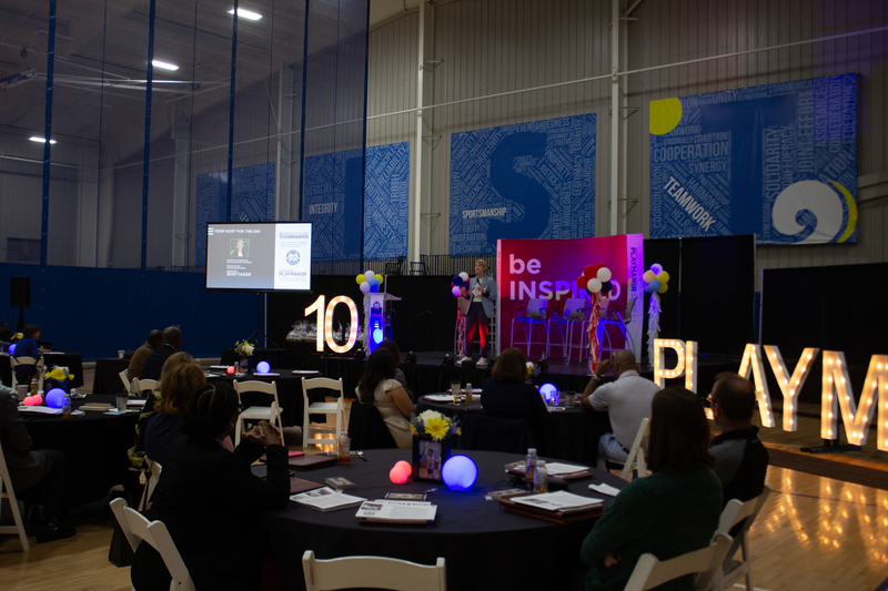 A conference room with a stage displaying "be inspired" in bright text. Attendees sit at round tables with decorative lights, under a backdrop of blue banners. The atmosphere is engaging and motivational.