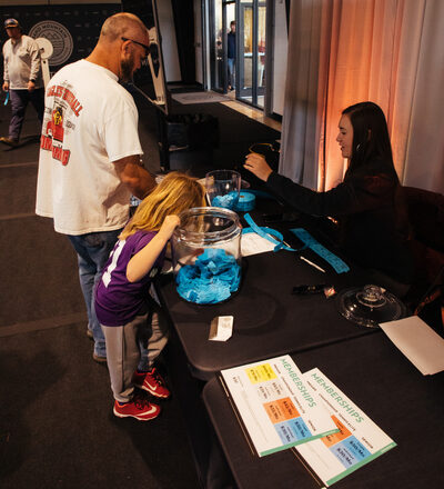 A man and a child stand at a table with a woman handing them raffle tickets. Two jars filled with blue tickets are on the table. Room has soft lighting.