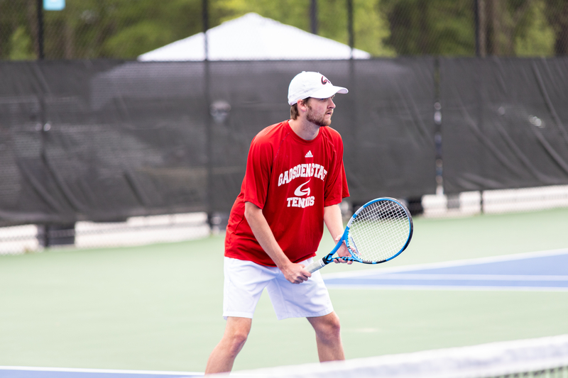 A tennis player in a red shirt and white shorts stands focused on a court, ready with a blue racket. The background shows a blurred fence and trees.