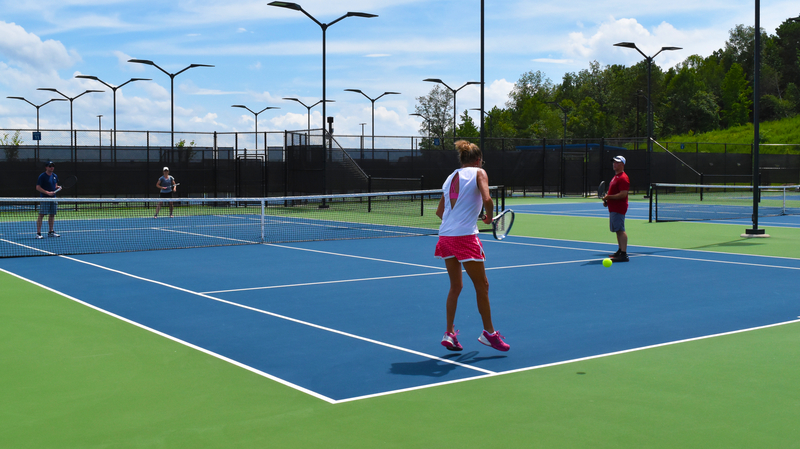 A group of four adults play doubles tennis on a sunny day. They're on a blue and green court, surrounded by trees and a clear sky. The mood is energetic.