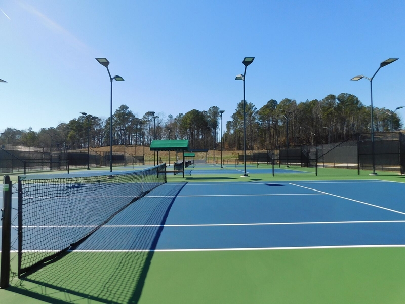 Two bright, blue and green tennis courts under a clear blue sky, surrounded by trees. The scene is calm, with overhead lights and nets.