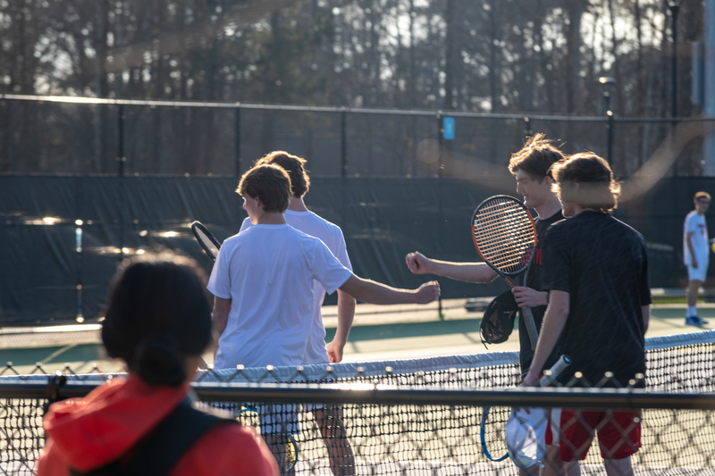 Teen tennis players exchange handshakes over the net post-match. Sunlight creates a warm glow, highlighting camaraderie on the court.